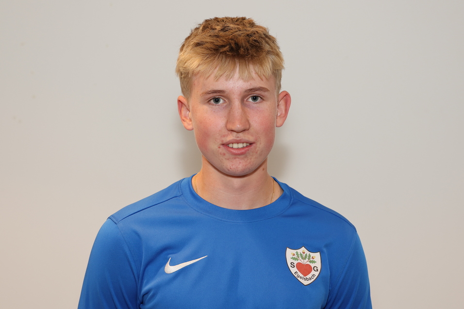 Junger Mann mit blondem Haar in blauer Sportjacke, l&auml;chelt vor grauem Hintergrund.