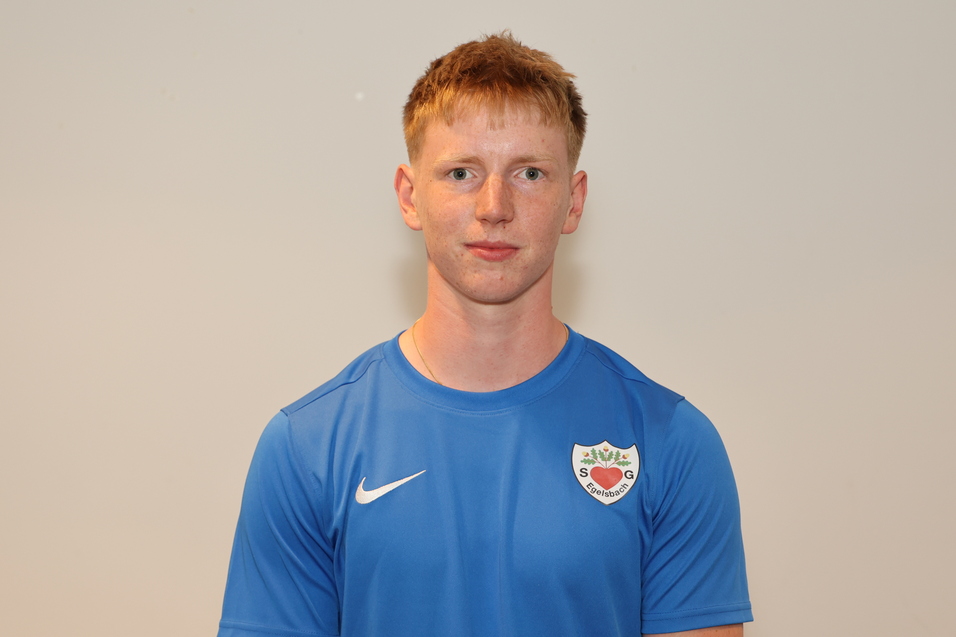 Ein junger Mann in blauem Sportshirt steht vor einer hellen Wand und schaut direkt in die Kamera.