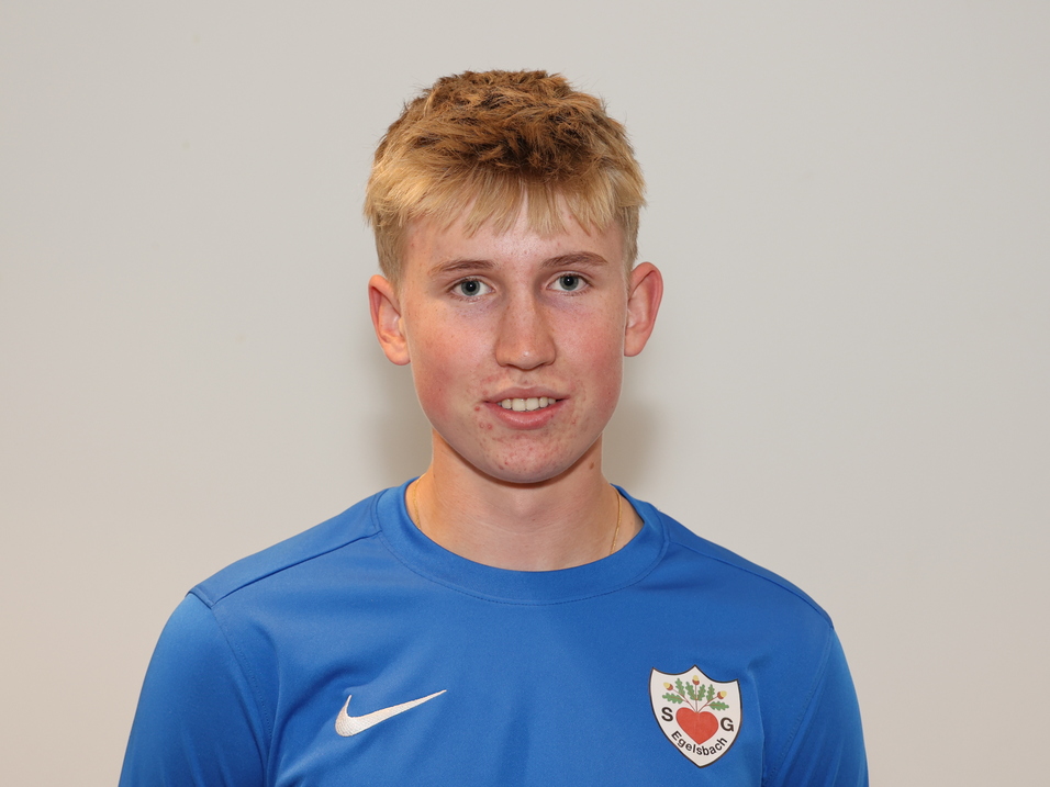 Junger Mann mit blondem Haar in blauer Sportjacke, l&auml;chelt vor grauem Hintergrund.