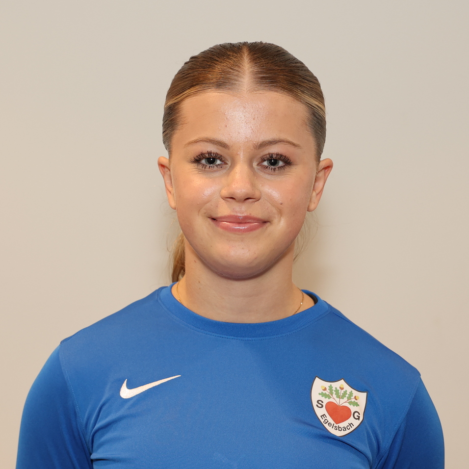 Portr&auml;t einer l&auml;chelnden jungen Frau in einem blauen Sportshirt mit Vereinswappen vor einem hellen Hintergrund.