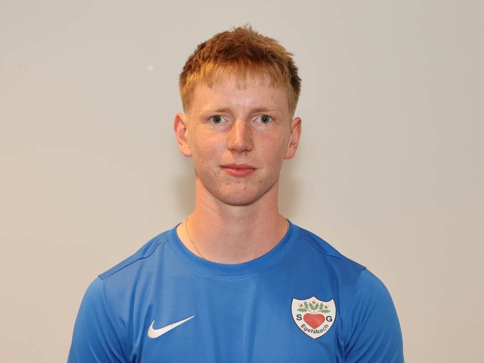 Ein junger Mann in blauem Sportshirt steht vor einer hellen Wand und schaut direkt in die Kamera.