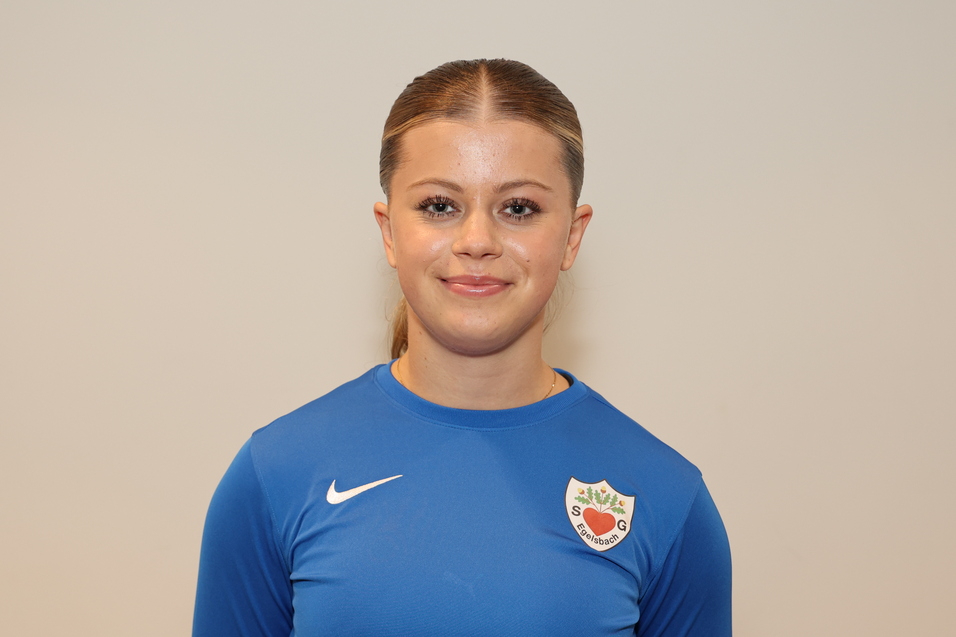 Portr&auml;t einer l&auml;chelnden jungen Frau in einem blauen Sportshirt mit Vereinswappen vor einem hellen Hintergrund.