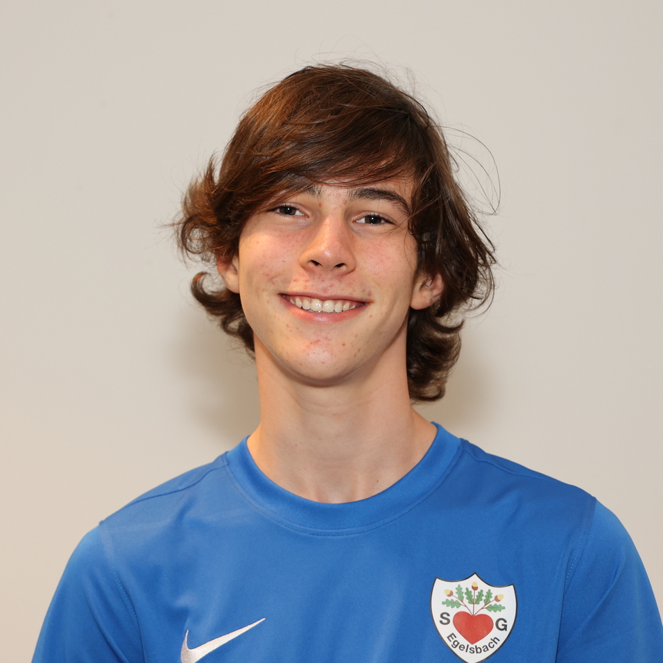 Junger Mann mit lockigem Haar tr&auml;gt ein blaues Fu&szlig;balltrikot und l&auml;chelt vor einem neutralen Hintergrund.