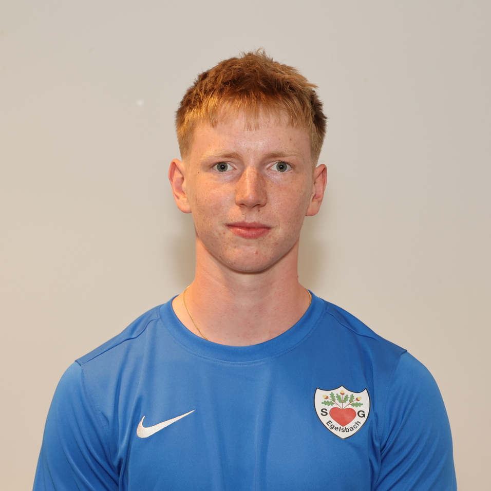 Ein junger Mann in blauem Sportshirt steht vor einer hellen Wand und schaut direkt in die Kamera.