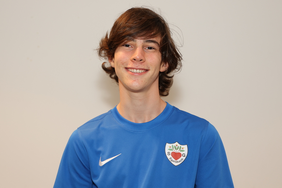Junger Mann mit lockigem Haar tr&auml;gt ein blaues Fu&szlig;balltrikot und l&auml;chelt vor einem neutralen Hintergrund.