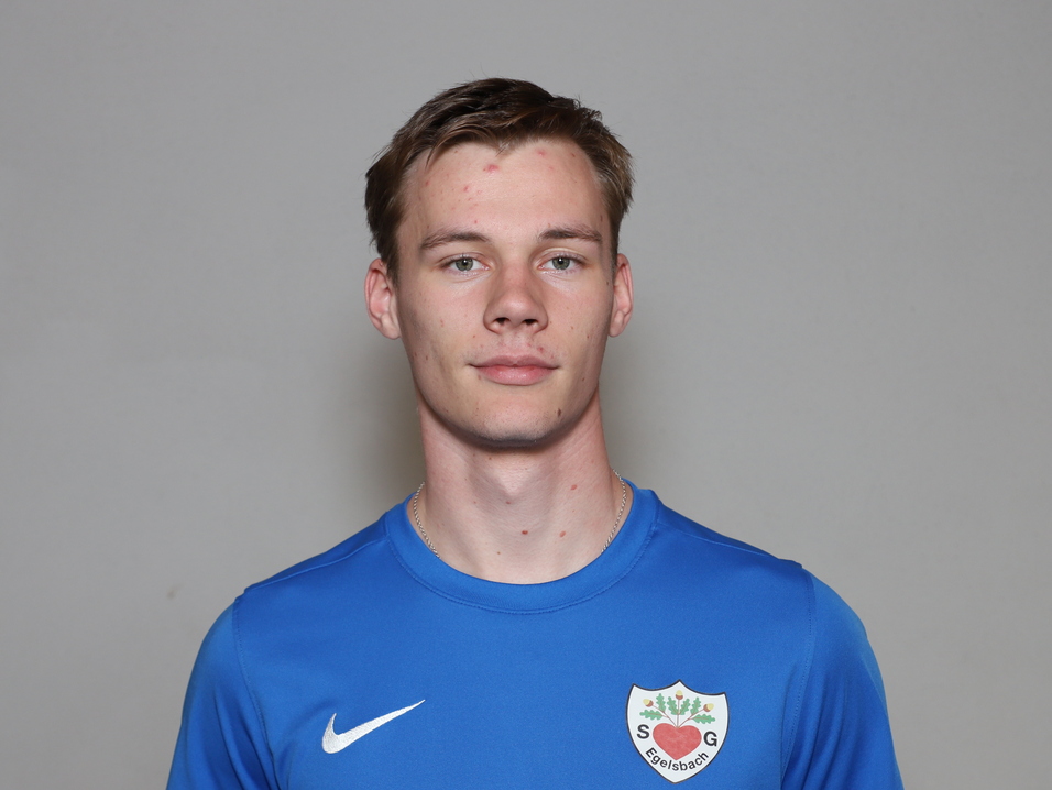 Junger Mann mit kurzem Haar, tr&auml;gt ein blaues Sportshirt und steht vor einem neutralen grauen Hintergrund.