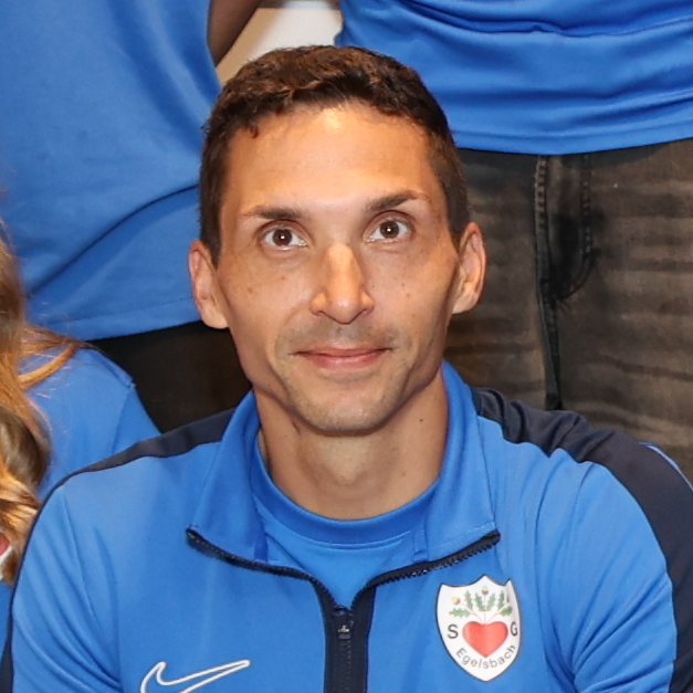 Gruppe von zw&ouml;lf Personen in blauen T-Shirts, die in zwei Reihen auf einem Fliesenboden stehen.