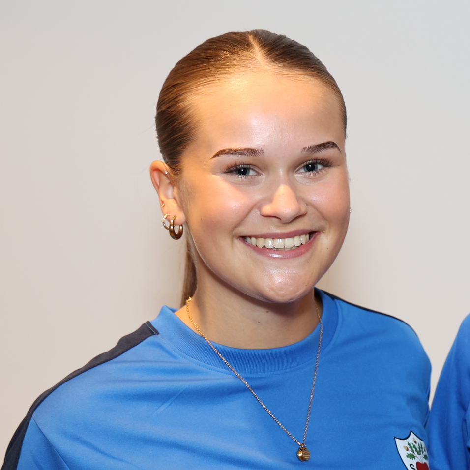 Zwei junge Frauen in blauen Sporttrikots stehen l&auml;chelnd nebeneinander vor einem hellen Hintergrund.