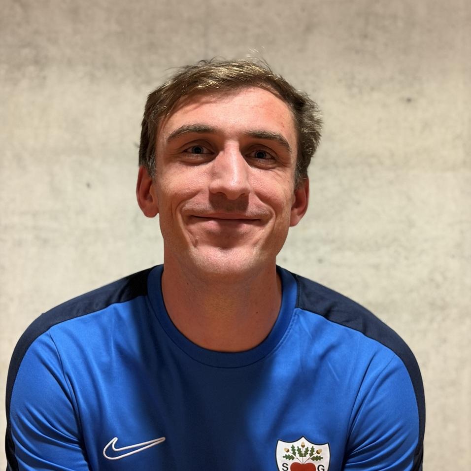 L&auml;chelnder Mann in blauer Sportbekleidung mit Vereinslogo vor einer grauen Wand.