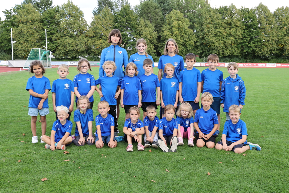 Gruppenfoto von 25 Kindern in blauen T-Shirts, umgeben von gr&uuml;ner Wiese und B&auml;umen im Hintergrund.