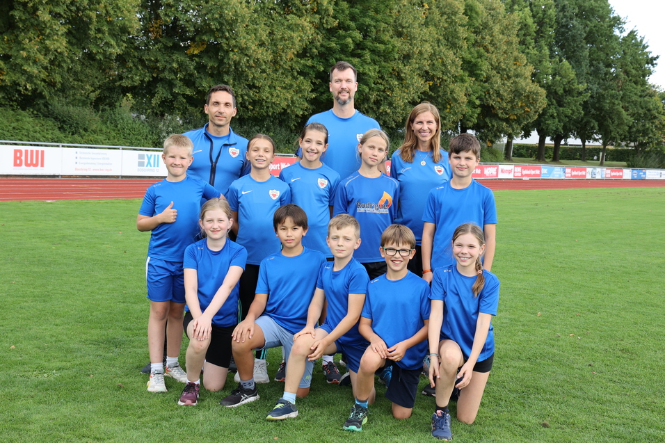 Gruppe von Kindern und Betreuern in blauen Sportshirts, l&auml;chelnd auf einer Laufbahn unter B&auml;umen.