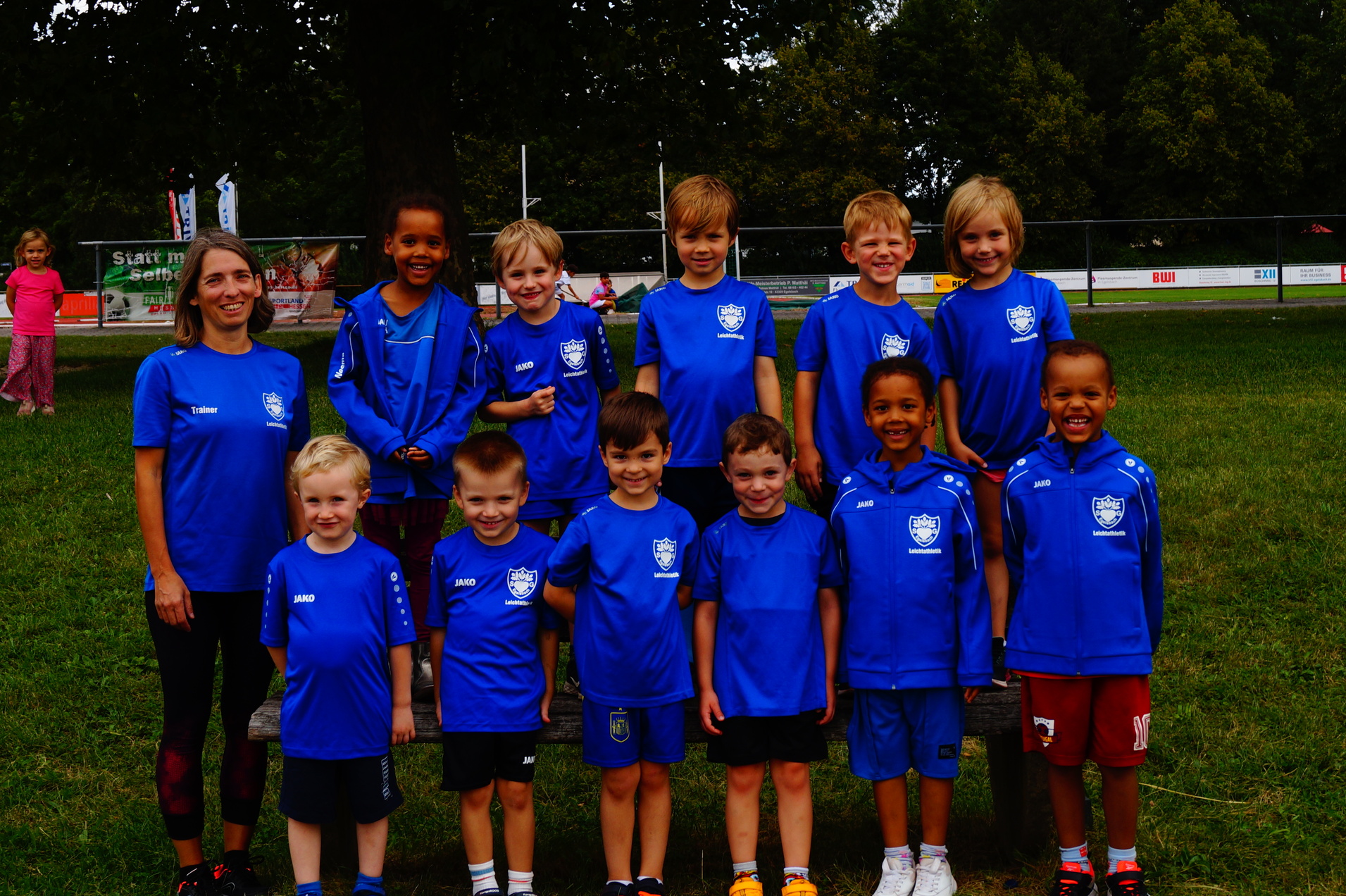 Gruppenfoto von zw&ouml;lf Kindern und einer Trainerin in blauen T-Shirts auf einem Sportplatz im Freien.
