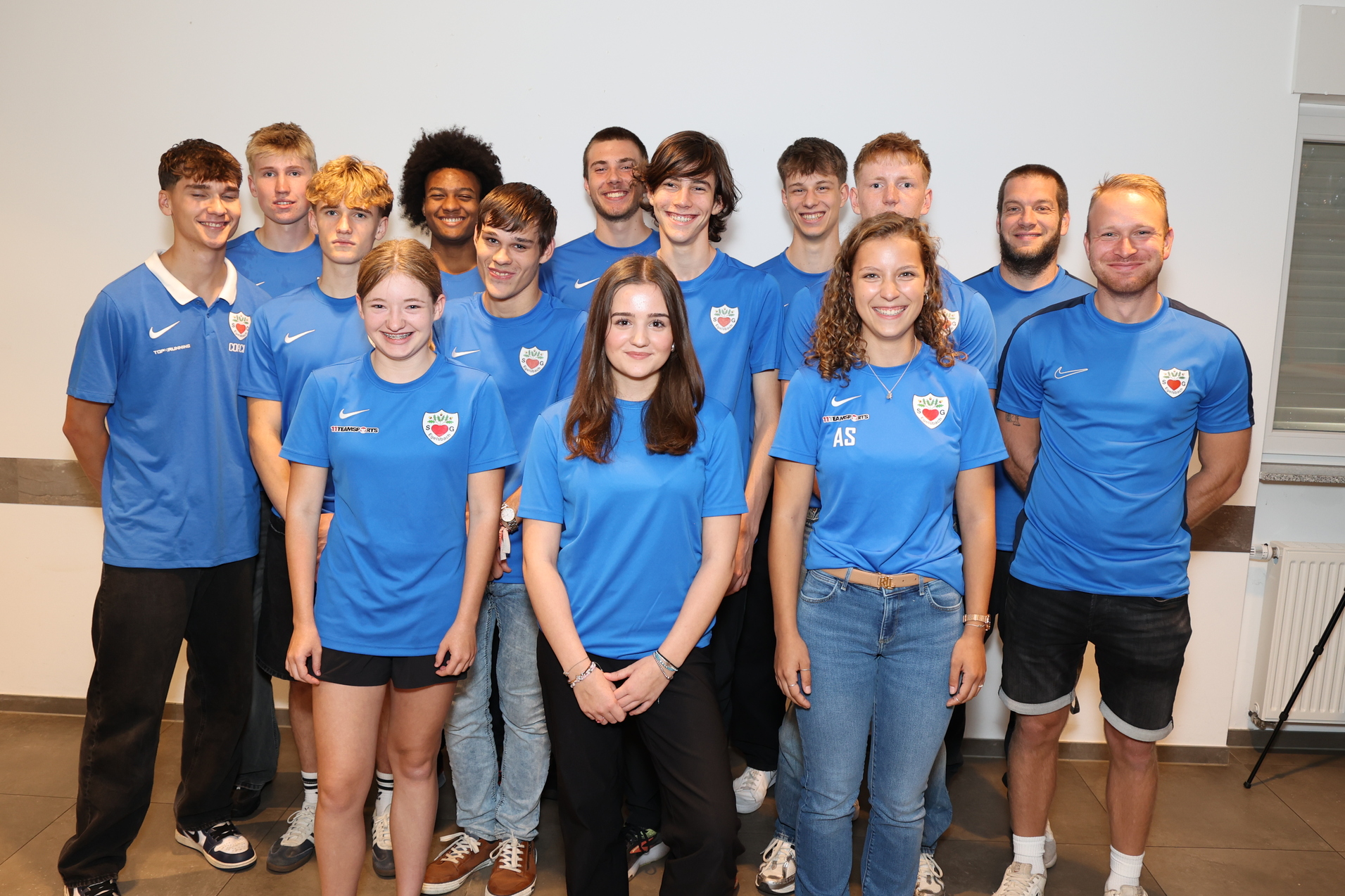 Gruppe von zw&ouml;lf Personen in blauen Sporttrikots, stehend in einem Raum mit neutralem Hintergrund.