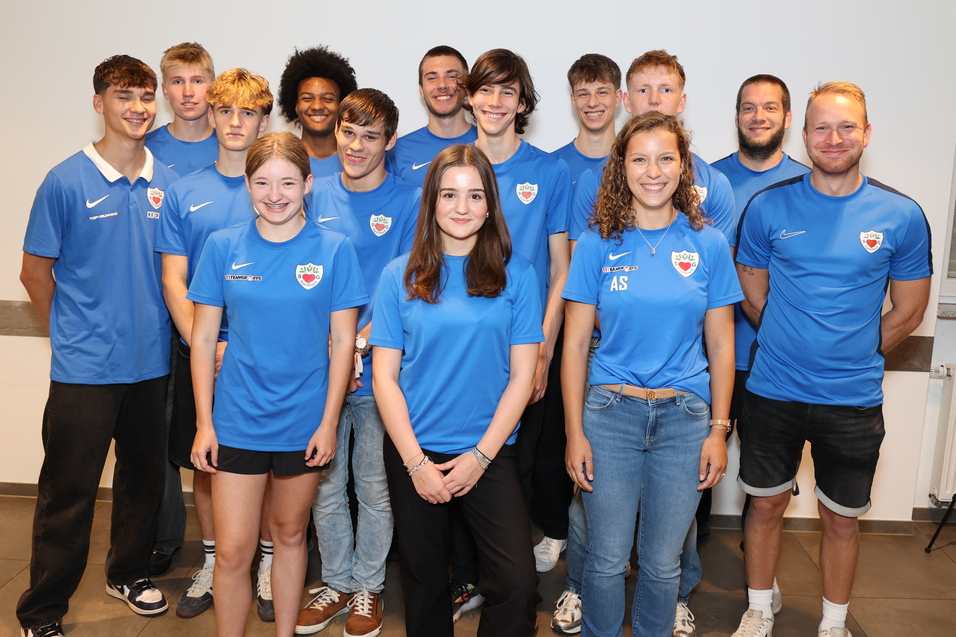 Gruppe von zw&ouml;lf Personen in blauen Sporttrikots, stehend in einem Raum mit neutralem Hintergrund.
