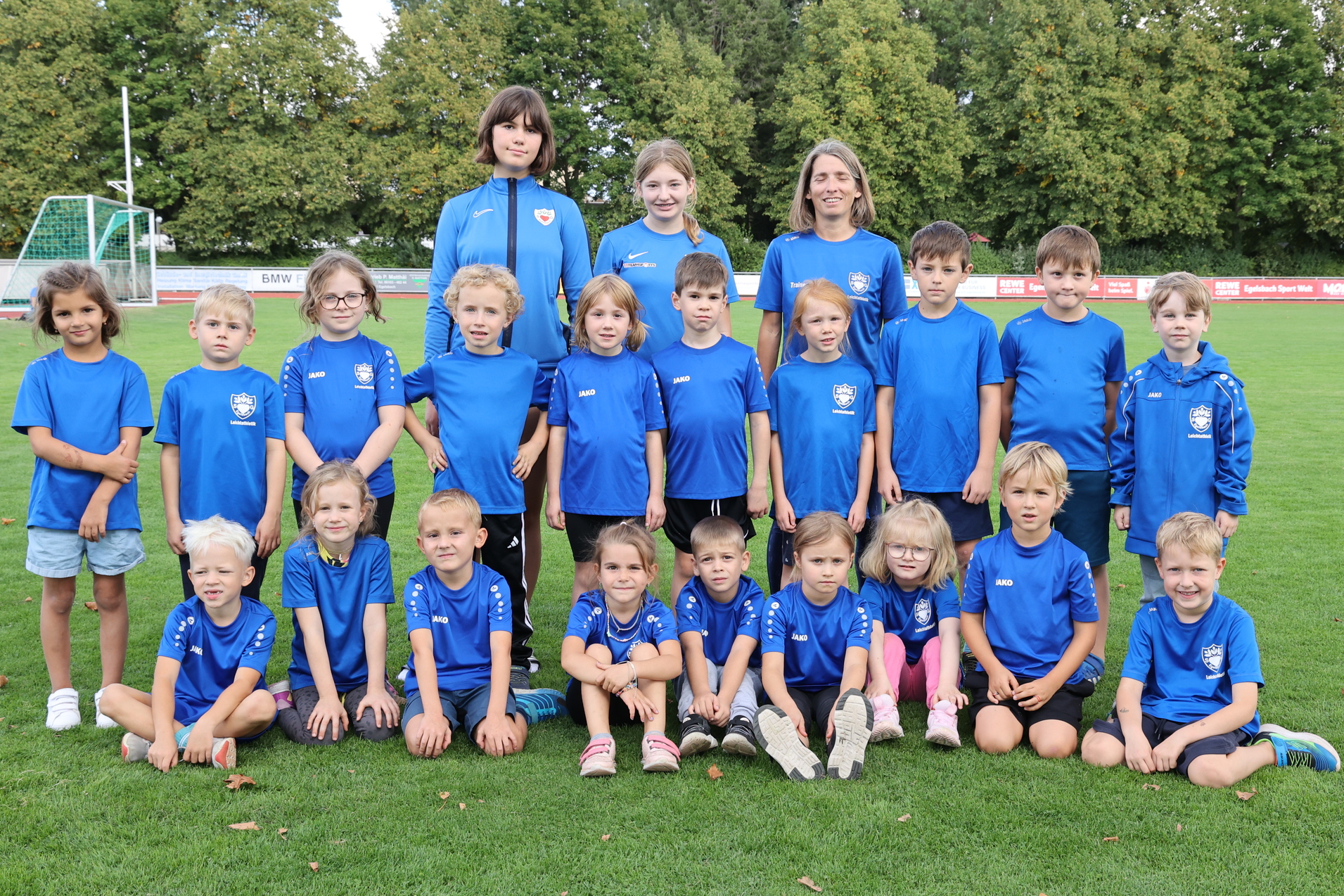 Gruppenfoto von 25 Kindern in blauen T-Shirts, umgeben von gr&uuml;ner Wiese und B&auml;umen im Hintergrund.