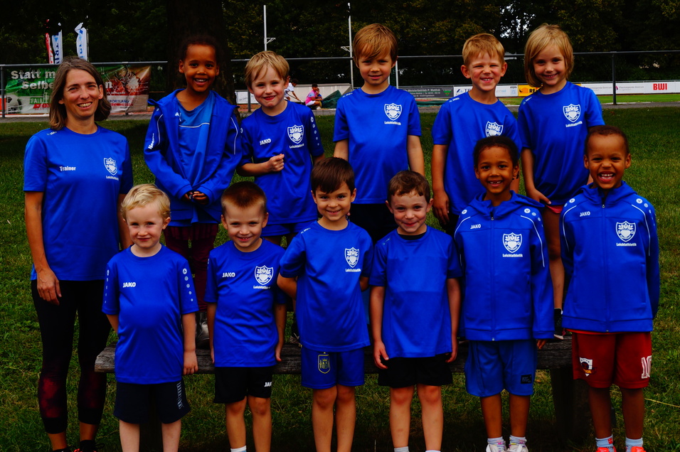 Gruppenfoto von zw&ouml;lf Kindern und einer Trainerin in blauen T-Shirts auf einem Sportplatz im Freien.