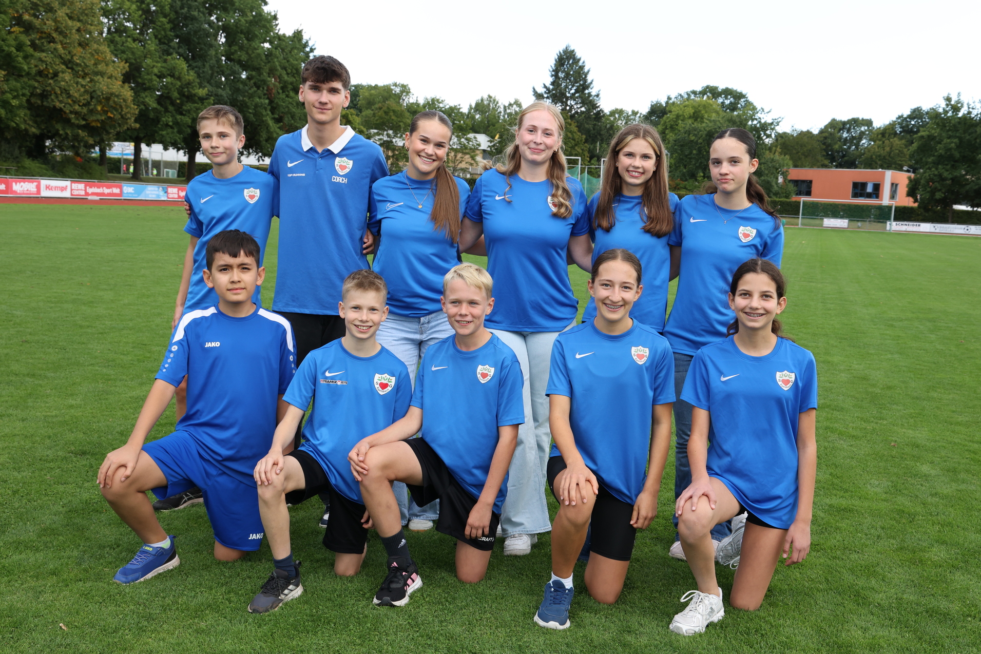 Gruppe von zehn Jugendlichen in blauen Sporttrikots, stehend und sitzend auf einem gr&uuml;nen Fu&szlig;ballfeld.