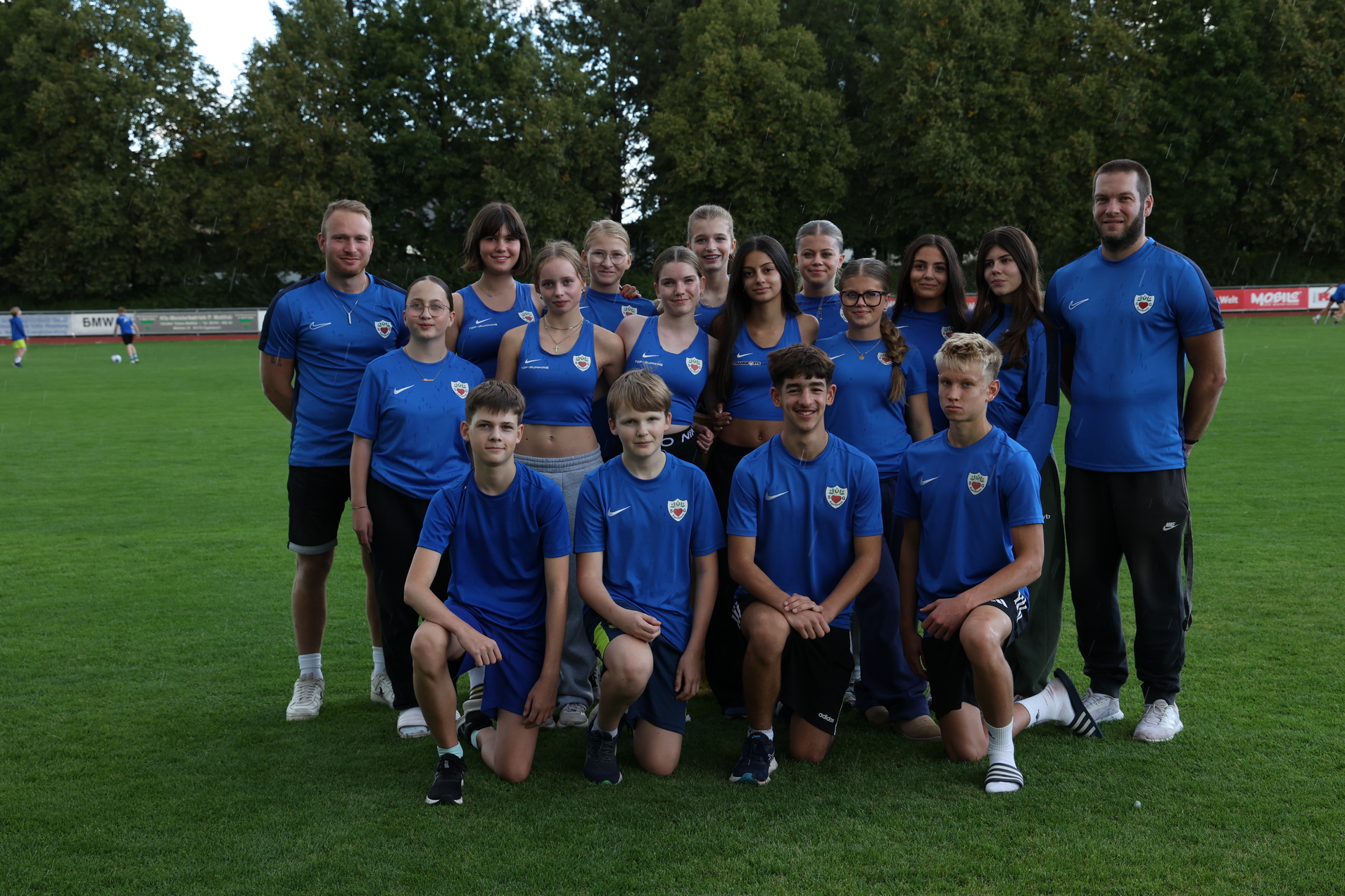 Gruppenshot von Jugendlichen und Trainern im blauen Trikot auf einem Rasenplatz, umgeben von B&auml;umen im Hintergrund.