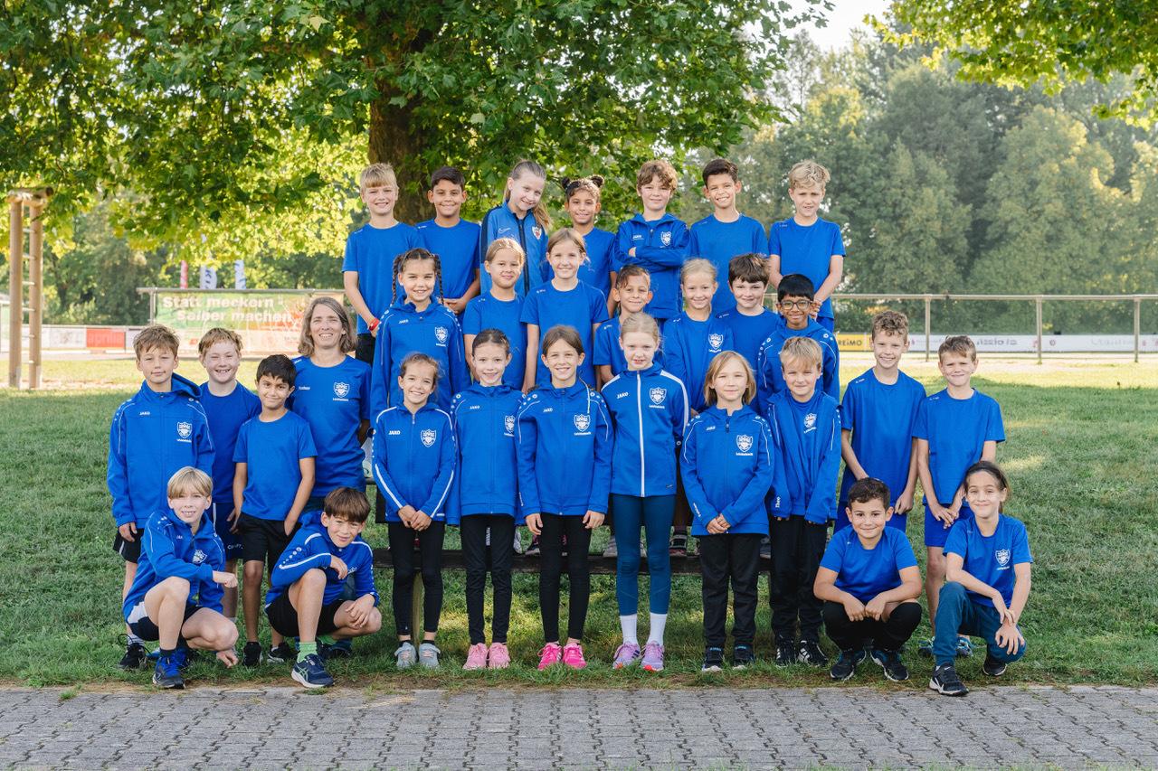 Gruppenfoto von 30 Kindern in blauen T-Shirts und Trainingsanzügen auf einer Wiese mit Bäumen im Hintergrund.