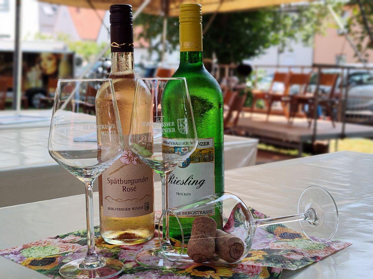 Zwei Weinflaschen und Gläser stehen auf einem Tisch mit einer bunten Unterlage und Korken daneben.