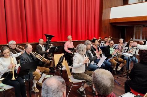 Blasorchester probt auf der B&uuml;hne mit verschiedenen Instrumenten vor rotem Vorhang. Dirigent leitet die Musiker.