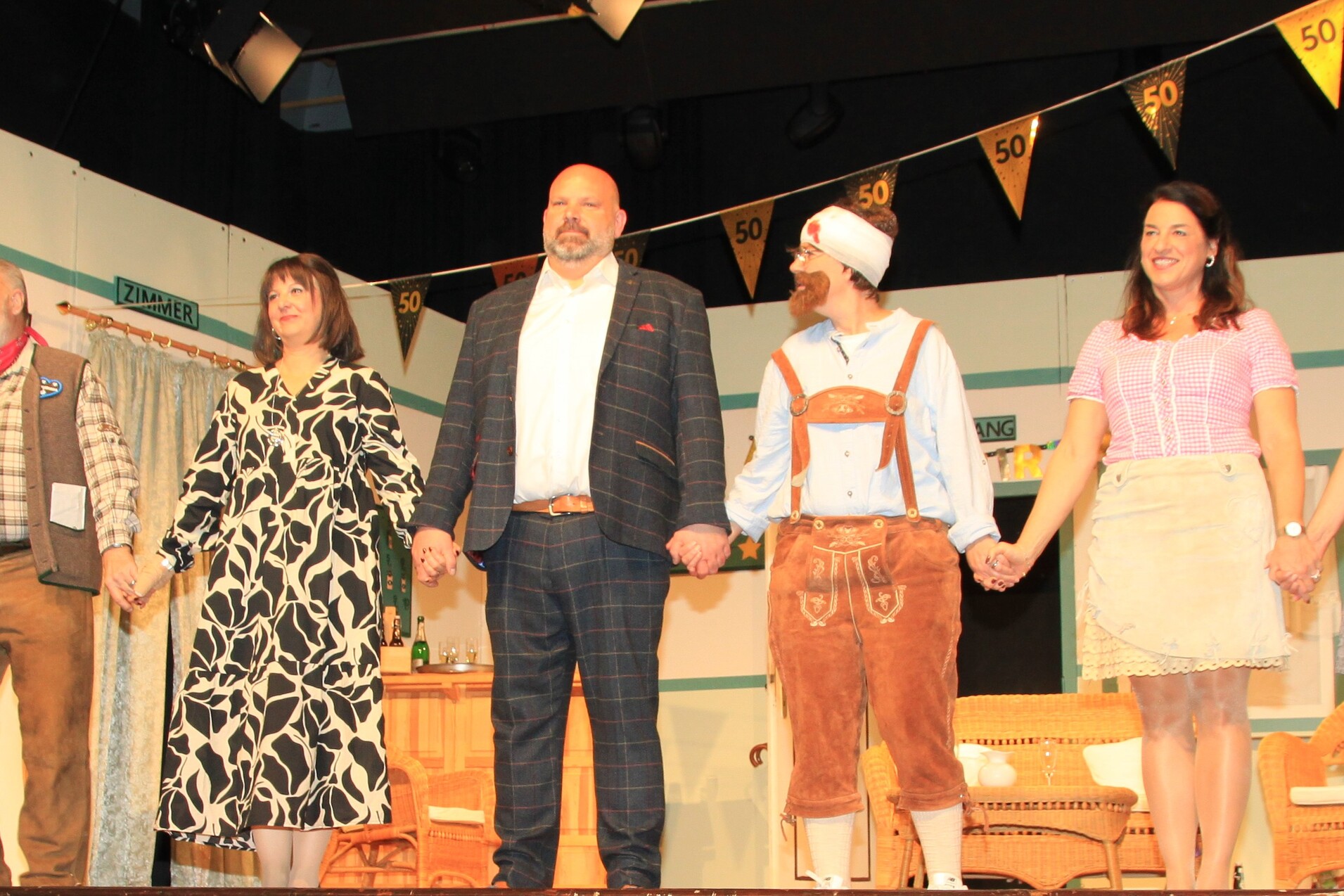 Acht Personen in festlicher Kleidung stehen auf einer B&uuml;hne und halten sich an den H&auml;nden, dekorierte Kulisse im Hintergrund.