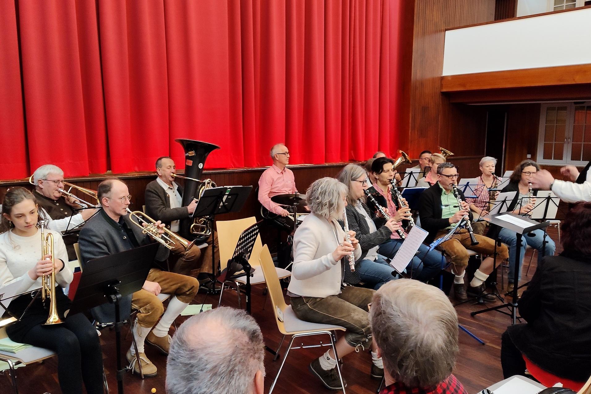 Blasorchester probt auf der B&uuml;hne mit verschiedenen Instrumenten vor rotem Vorhang. Dirigent leitet die Musiker.