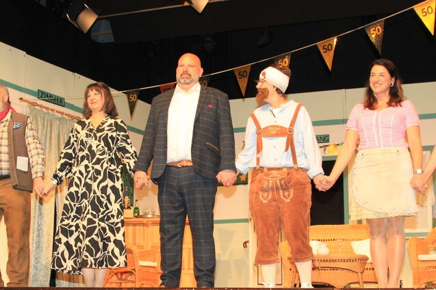 Acht Personen in festlicher Kleidung stehen auf einer B&uuml;hne und halten sich an den H&auml;nden, dekorierte Kulisse im Hintergrund.