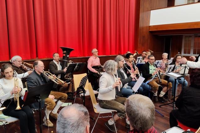 Blasorchester probt auf der B&uuml;hne mit verschiedenen Instrumenten vor rotem Vorhang. Dirigent leitet die Musiker.