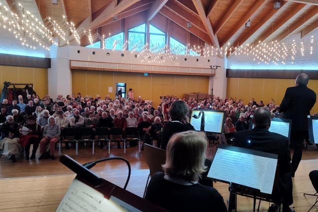 Blick auf ein Publikum bei einem Konzert, im Vordergrund Musiker und Notenst&auml;nder, hinter ihnen eine gef&uuml;llte Halle.