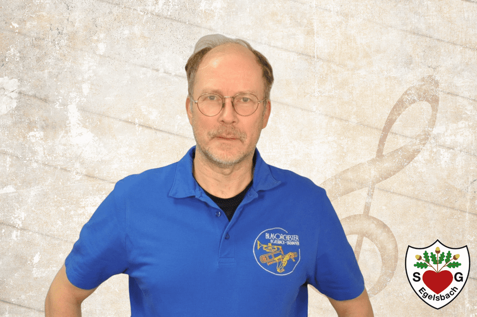 Mann mit Brille tragt ein blaues T-Shirt mit Logo vor einer strukturierten, neutralen Hintergrund.