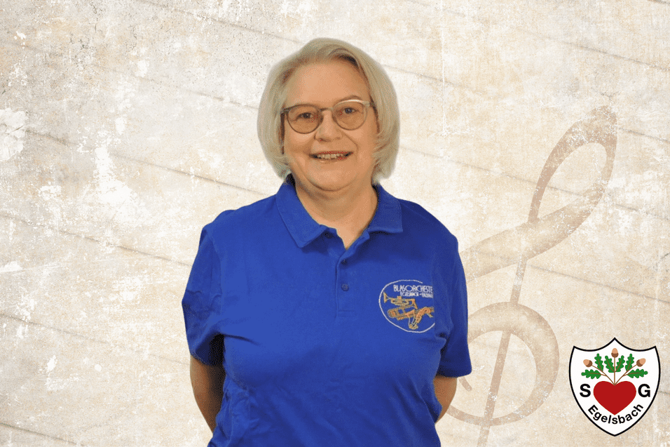 Frau mit blonden Haaren und Brille in blauer Bluse, stehend vor einem hellen, musikalischen Hintergrund.