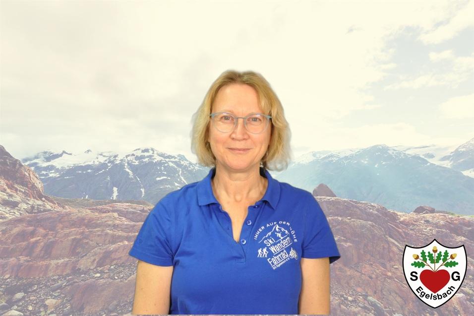 Porträt einer lächelnden Frau in einem blauen Shirt, im Hintergrund eine Gebirgslandschaft mit Wolken.