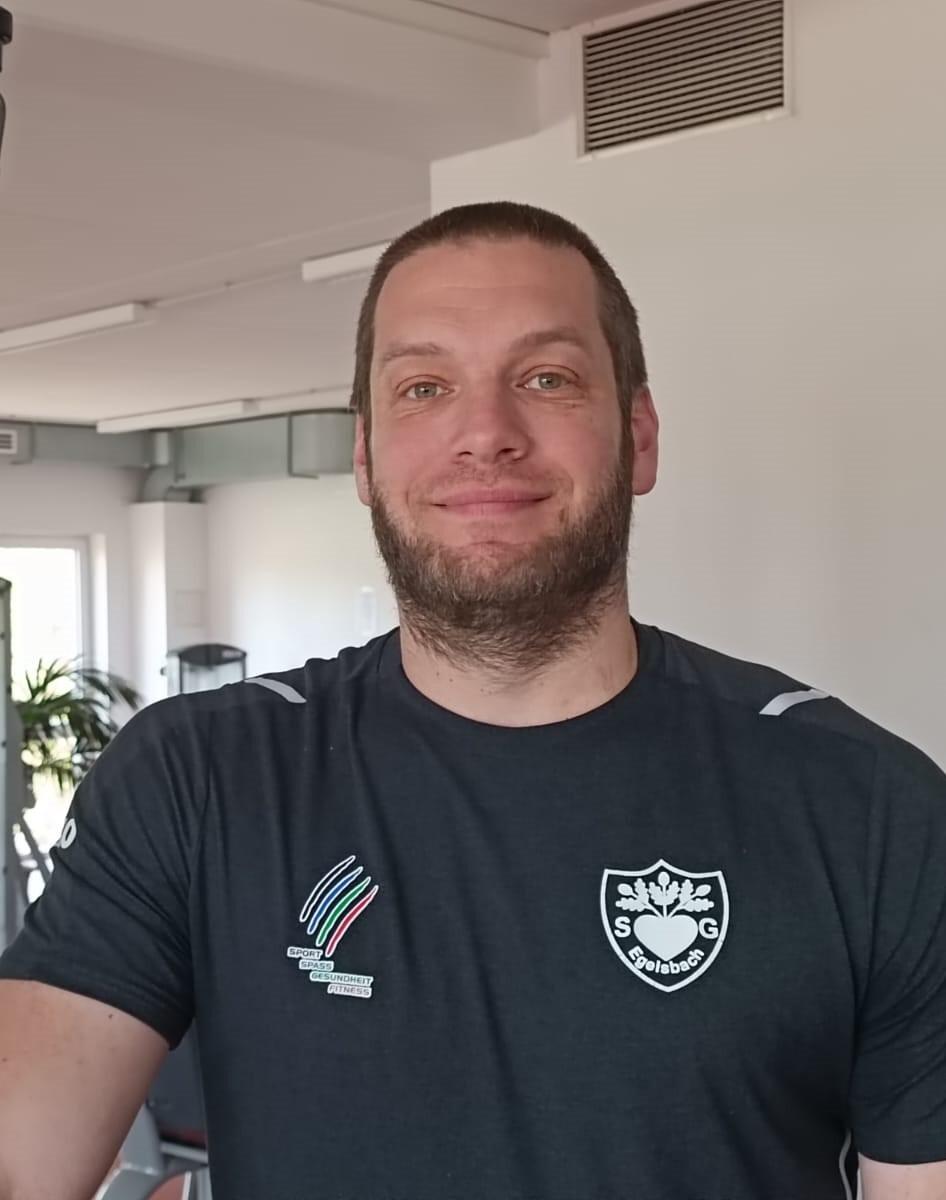 Mann mit Bart in Sportkleidung lächelt in einem Fitnessstudio mit modernem Ambiente.