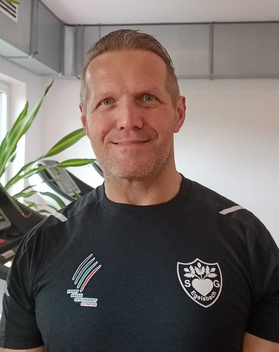 Mann mit kurzem Haar steht vor einem Fitnessgerät in einem hellen Raum, trägt ein schwarzes Shirt mit Logo.