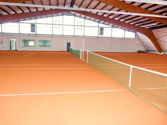 Innenansicht einer Tennishalle mit zwei Tennispl&auml;tzen und Netzen auf einem roten Belag unter einem sichtbaren Dach.