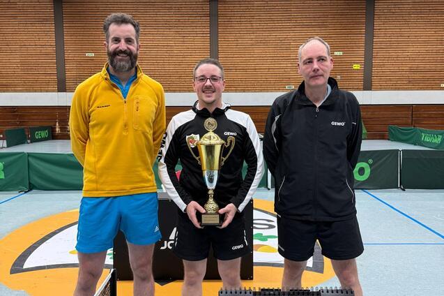 Drei Tischtennisspieler posing mit einem Pokal, im Hintergrund eine Tischtennisplatte und die Spielstandsanzeige.
