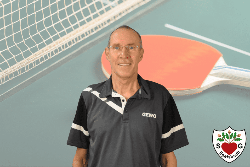 Porträt eines Mannes in einem schwarzen Tischtennisshirt vor einer Tischtennisplatte mit Schläger und Netz im Hintergrund.