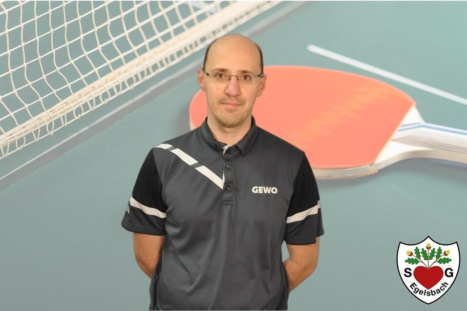 Mann in schwarzer Sportbekleidung steht vor einer Tischtennisplatte und einem Tischtennisschläger.