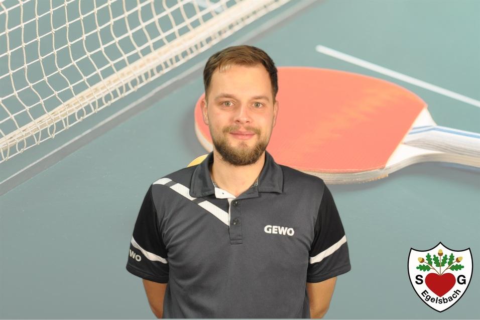 Portrait eines Mannes in einem Sportshirt vor einer Tischtennisplatte mit Schläger und Netz im Hintergrund.
