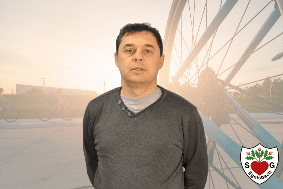 Mann in grauem Pullover steht vor verschwommenem Hintergrund mit Fahrr&auml;dern und Sonnenuntergang. Logo einer Sportgemeinschaft in der Ecke.