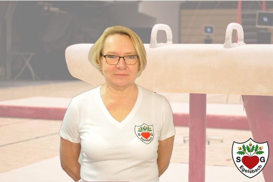 Frau mit Brille in weißem T-Shirt steht vor einem Turngerät in einer Sporthalle.