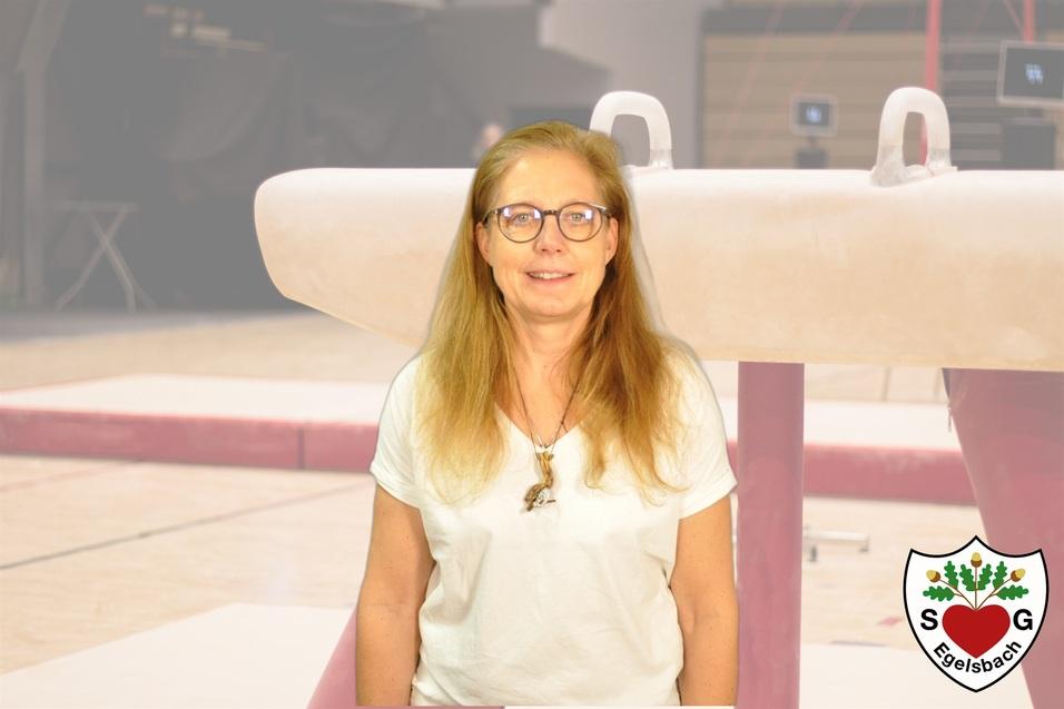 Frau mit langen Haaren und Brille steht lächelnd vor einem Gerätturngerät in einer Halle.