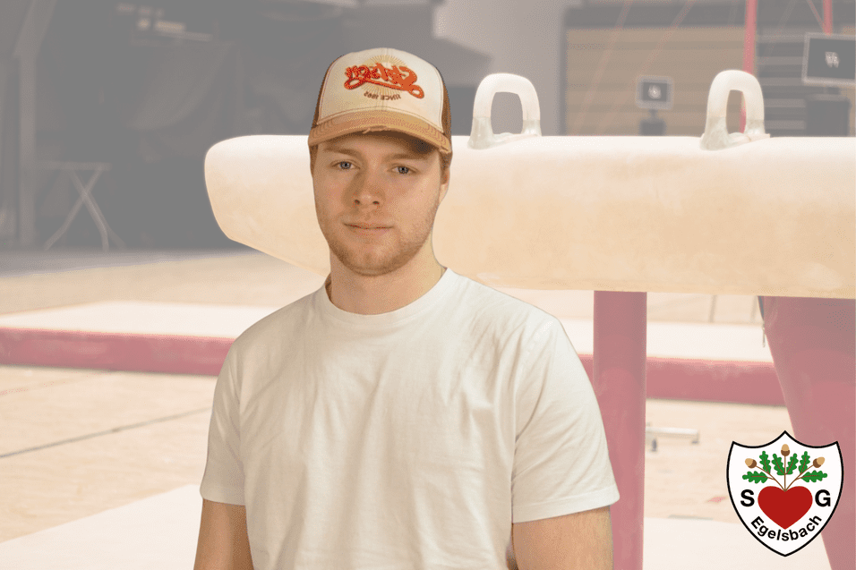 Junger Mann in weißem Shirt steht vor einem Barren in einer Sporthalle mit leicht verschwommenem Hintergrund.