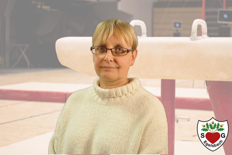 Frau mit blonden Haaren und Brille steht vor einem Gerätepark in einer Turnhalle mit einem Barren im Hintergrund.