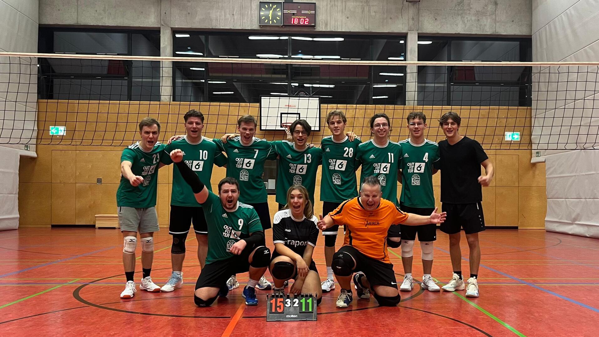 Volleyballmannschaft posiert nach dem Spiel, alle in gr&uuml;nen Trikots, mit Ergebnistafel 15:11 auf dem Boden.