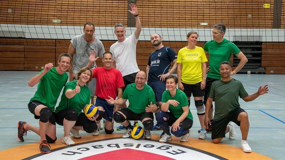 Gruppe von Volleyballspielern in bunter Sportkleidung posiert l&auml;chelnd auf dem Spielfeld vor einem Netz.