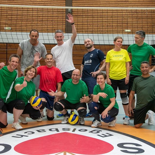 Gruppe von Volleyballspielern in bunter Sportkleidung posiert l&auml;chelnd auf dem Spielfeld vor einem Netz.