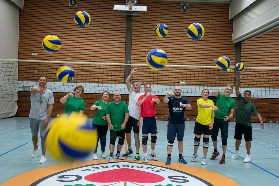 Gruppe von zehn Personen in Sportbekleidung hebt Volleyballs in einer Halle mit Netz und blauer Sportfläche.