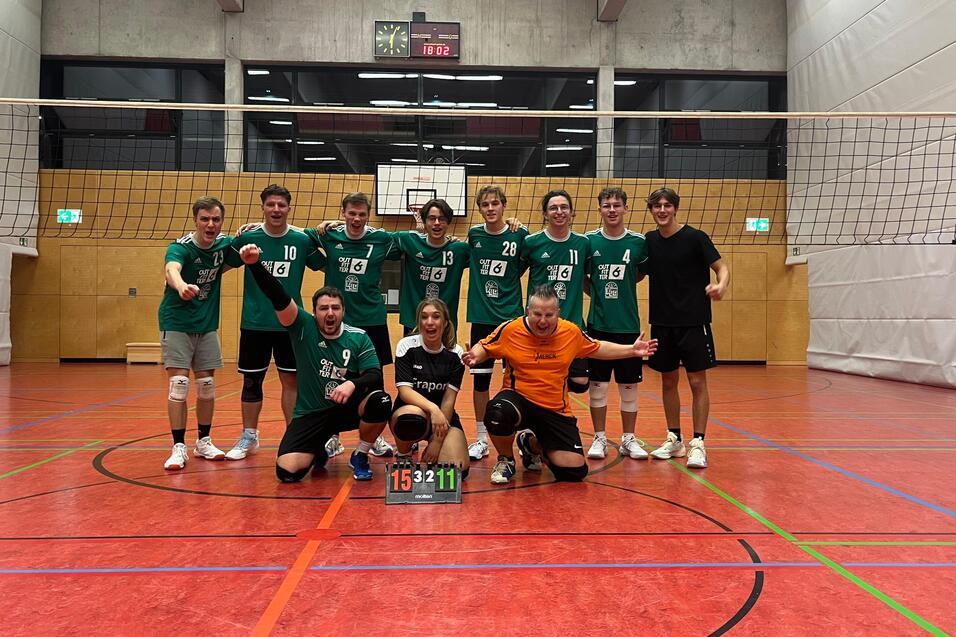 Volleyballmannschaft posiert nach dem Spiel, alle in grünen Trikots, mit Ergebnistafel 15:11 auf dem Boden.